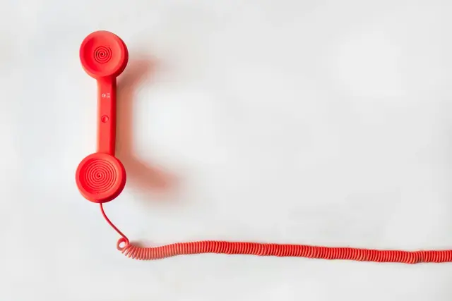 A vibrant red, retro telephone handset rests on a stark white background, accompanied by a long, matching coiled cord. The minimalist composition creates a clean, bold aesthetic that highlights the classic design of the communication device.