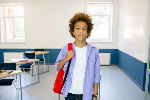 A young boy with curly hair stands in a clean, modern classroom, wearing a white t-shirt, a lavender button-down shirt, and a bright red backpack. He looks directly at the camera with a poised and calm expression.
