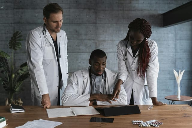 Three healthcare professionals in white lab coats and stethoscopes gather around a wooden table in a modern office, collaboratively reviewing information on a tablet. The mood is focused and professional, highlighted by a clean, minimalist workspace that features medical records, loose documents, and an assortment of medication blister packs.