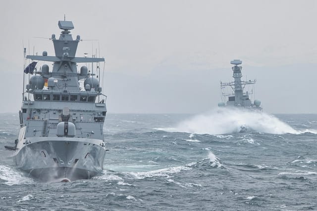 Two modern warships navigate through turbulent, choppy ocean waters under a gray, overcast sky, demonstrating maritime strength in challenging conditions. The lead ship, adorned with a German flag, cuts through the waves while the second vessel follows behind, creating a powerful sense of naval cooperation.