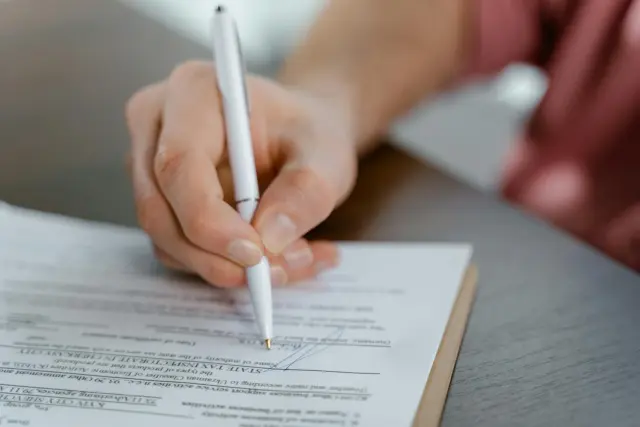 A close-up shot captures a person's hand signing an official Ukrainian document with a white ballpoint pen, conveying a professional and focused atmosphere. The depth of field is shallow, drawing sharp attention to the act of signing while softly blurring the surrounding workspace.