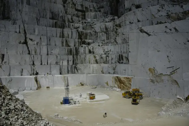 A vast marble quarry features towering, vertically cut white stone walls overlooking a wide, pale floor where heavy yellow machinery and small figures of workers are stationed. The scene conveys a sense of industrial scale and stark, monumental contrast between the massive, rugged rock faces and the human-operated equipment below.