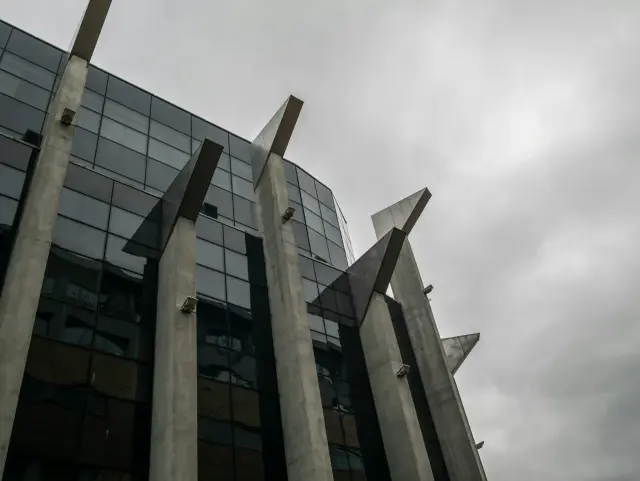 The image depicts a low-angle view of a modern, glass-paneled office building characterized by prominent, wing-like concrete structural supports protruding from its facade against a somber, overcast sky. The architectural composition creates a stark, geometric aesthetic with a moody and somewhat industrial atmosphere.