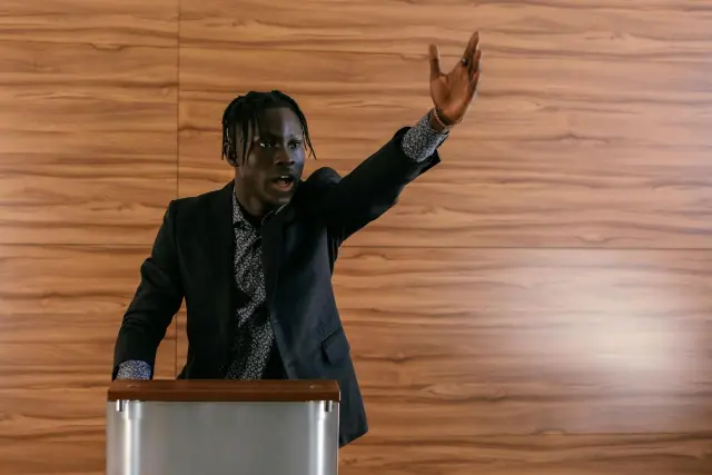 A young man with braided hair, dressed in a sharp blazer and patterned shirt, gestures expressively while speaking passionately behind a metallic podium. He stands against a modern, warm-toned wood-paneled wall, conveying a mood of intense focus and authoritative engagement.