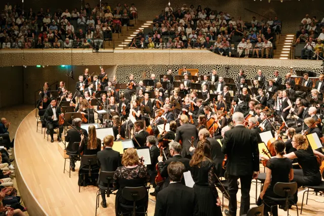 A formal orchestra performs on stage in a modern, architecturally distinctive concert hall, with a large audience watching from the tiered seating above. The scene captures a professional and disciplined atmosphere, highlighting the contrast between the dark attire of the musicians and the warm, wood-toned interior of the venue.