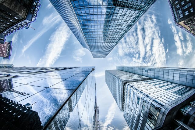 A low-angle, wide-lens perspective captures towering modern skyscrapers in Frankfurt, their glass facades reflecting the vast, cloud-streaked blue sky. The dramatic upward composition emphasizes the scale and geometric complexity of the urban architecture, creating an imposing and crisp atmosphere.