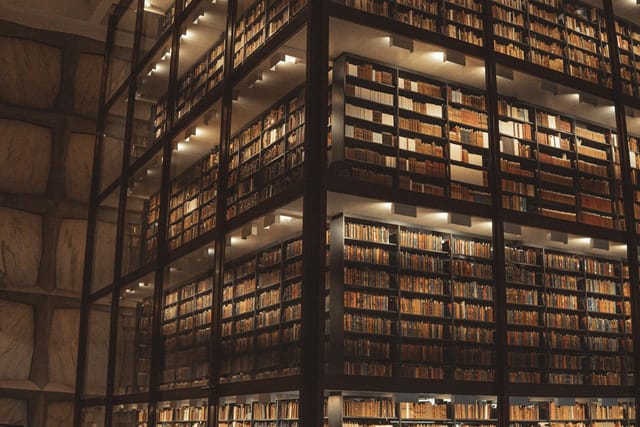 This image depicts the dramatic, multi-story glass-enclosed bookshelves of Yale University’s Beinecke Rare Book and Manuscript Library, creating an awe-inspiring and scholarly atmosphere. The warm, focused lighting illuminates the countless volumes, emphasizing the grand, grid-like scale of the unique architectural structure.