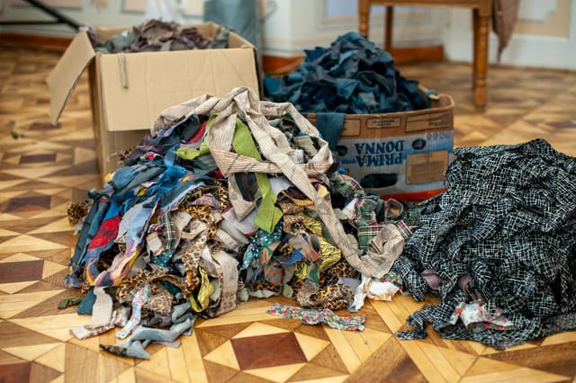 This image features several large, disorganized piles of fabric scraps—ranging from patterned prints to solid colors—resting on a classic parquet wooden floor. The scene has a resourceful, creative, and somewhat chaotic atmosphere, highlighted by the use of cardboard boxes for storage in a bright, indoor workspace.